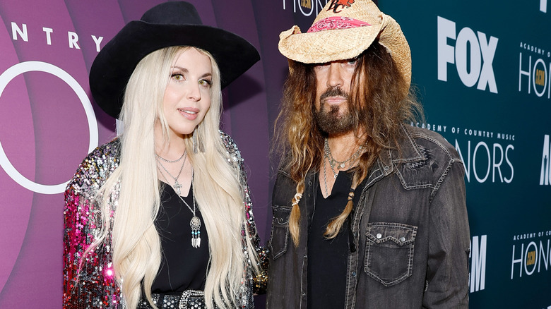 Billy Ray Cyrus in cowboy hat and denim shirt with long hair and Firerose Cyrus in black hat and black ripped jeans