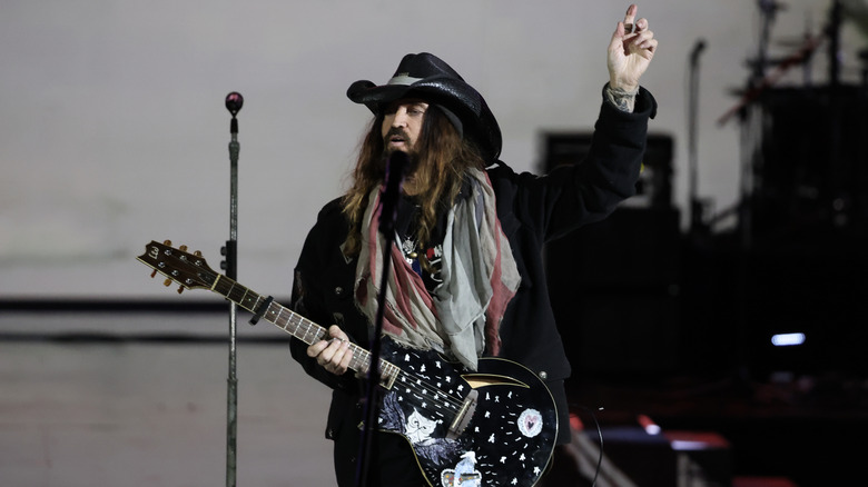 Billy Ray Cyrus in black hat and gray scarf holding guitar in front on mic stand