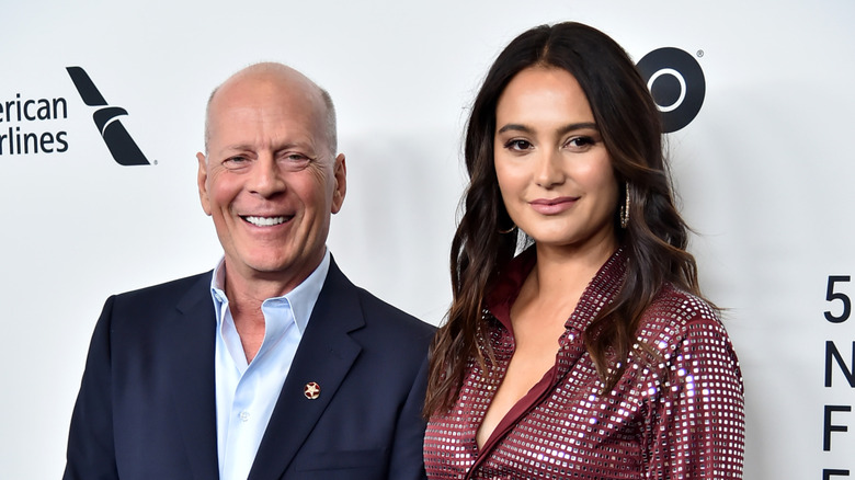 Bruce Willis smiling in a suit next to Emma Heming who's wearing a red dress.