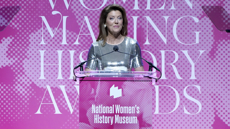 Norah O'Donnell standing behind a clear podium in front of a pink sign.