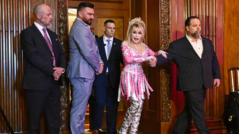 Dolly Parton in a strappy pink dress holds on to her security man as she walks into a wood paneled room