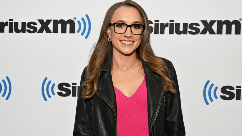 Kat Timpf smiling in glasses, a black leather jacket, and a pink sweater