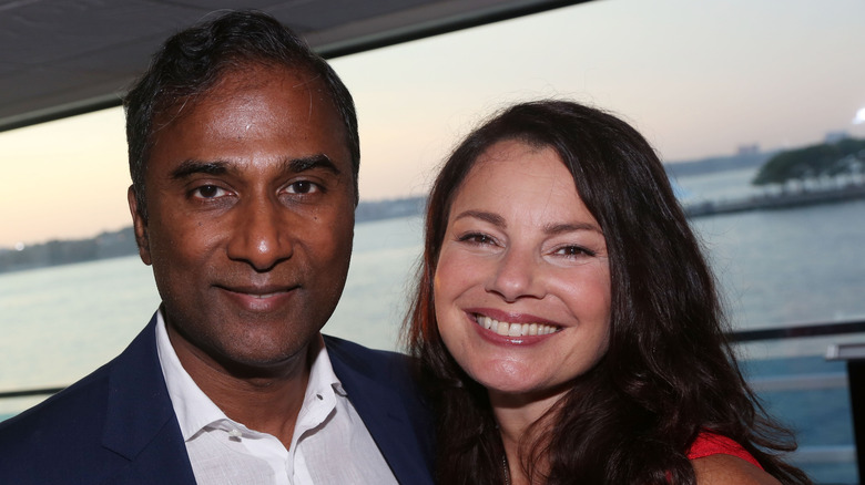 Fran Drescher smiling in a red dress next to Shiva Ayyadurai in a blazer and white collared shirt while they pose in front of an ocean.