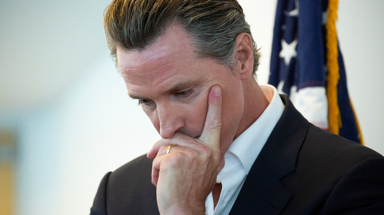 Gavin Newsom looking deep in thought with his finger on his cheek in a dark suit