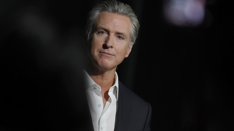 Gavin Newsom looking thoughtful in a dark suit with a white open-collared shirt