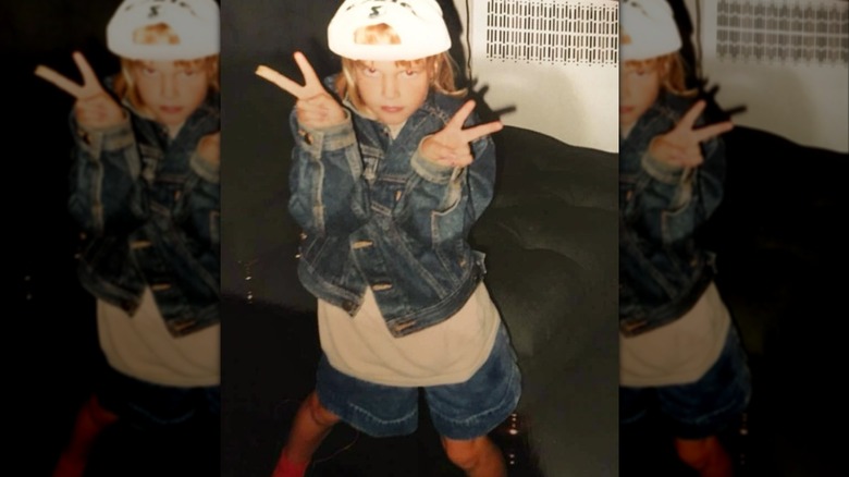 Young Madeline Brewer in backwards ball cap, jean jacket, and shorts making peace signs