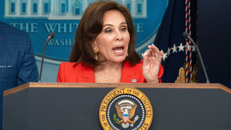Jeanine Pirro delivering a speech at a podium in a red blazer with an American flag pin