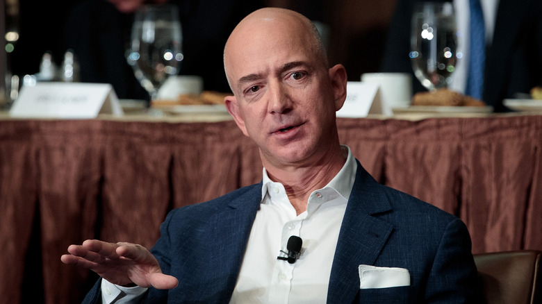 Jeff Bezos speaking while raising his hand in a navy jacket and white shirt