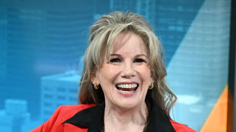 Melissa Gilbert smiling and resting her arm on the back of the couch she's sitting on in a red blazer