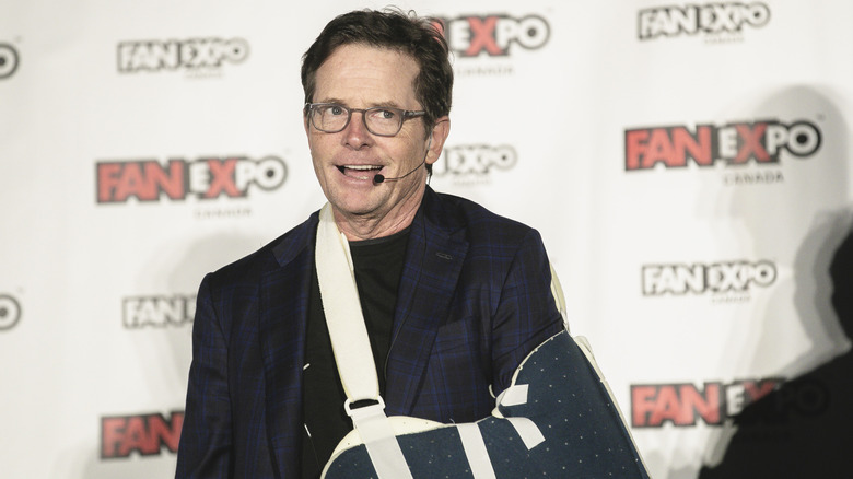 Michael J. Fox smiling with his arm in a sling in glasses and a navy blazer