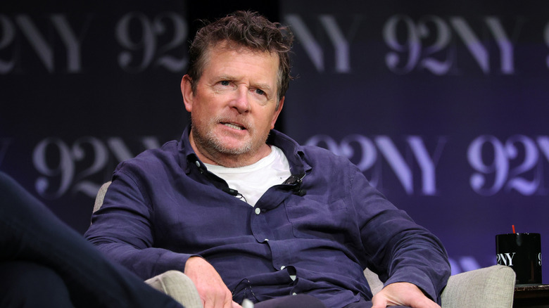 Michael J. Fox making a face while sitting onstage in a navy shirt with a white T-shirt underneath