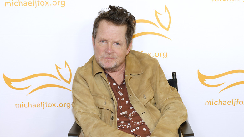 Michael J. Fox posing for photos in a beige jacket with a brown patterned shirt underneath