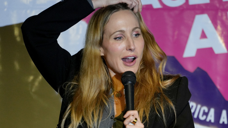 Nikki Glaser talking into microphone and touching the top of her head