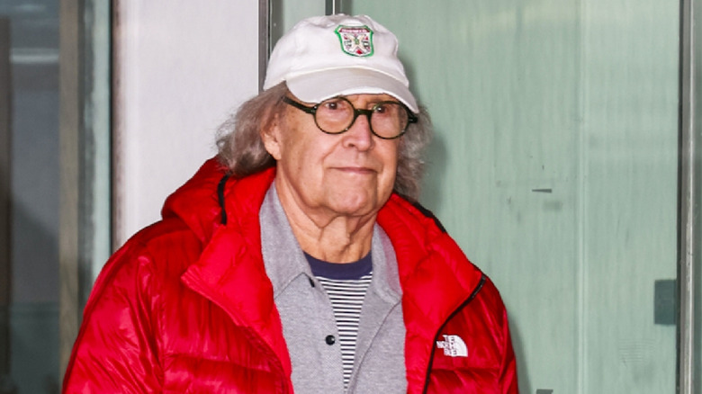 Chevy Chase in white ball cap, glasses, and red jacket with long gray hair
