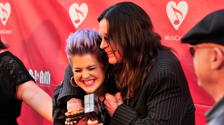 Kelly Osbourne with short purple hair being hugged by father Ozzy in suit and sunglasses