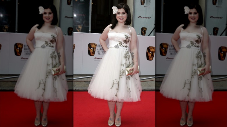 Kelly Osbourne on a red carpet in white dress with white flower in her black hair