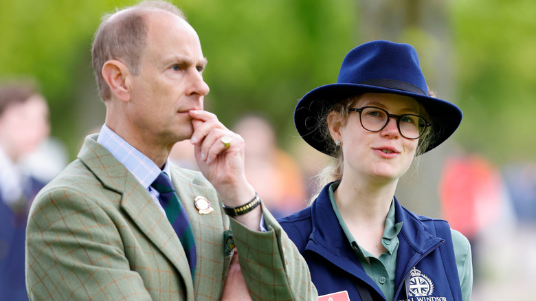 Prince Edward with finger to his lip next to Lady Louise Mountbatten-Windsor in a blue hat