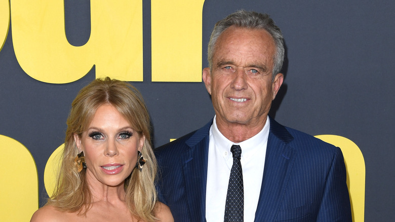 Cheryl Hines wearing an uncomfortable smile in a red dress next to RFK Jr. in a navy suit