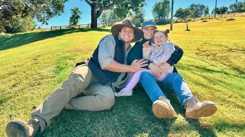 Chandler Powell, Bindi Irwin, and their daughter Grace Warrior smile while posing for a photo on a grassy field in June 2025 (2025)
