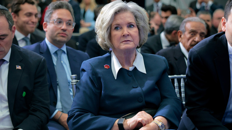 Susie Wiles looking serious while seated at an event in a navy suit jacket with a white shirt underneath
