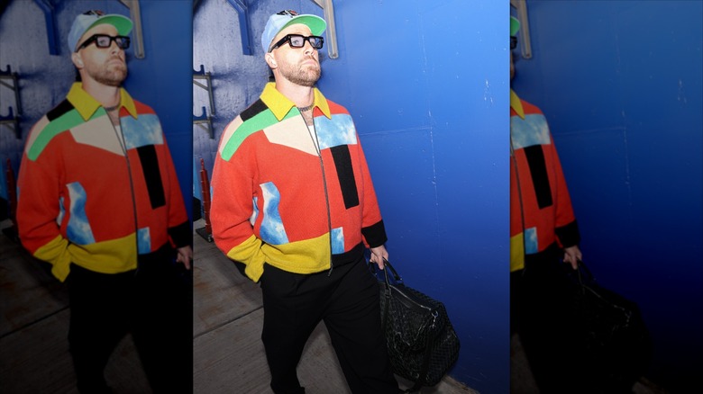 Travis Kelce with a baseball hat and glasses walking in a blue hallway.