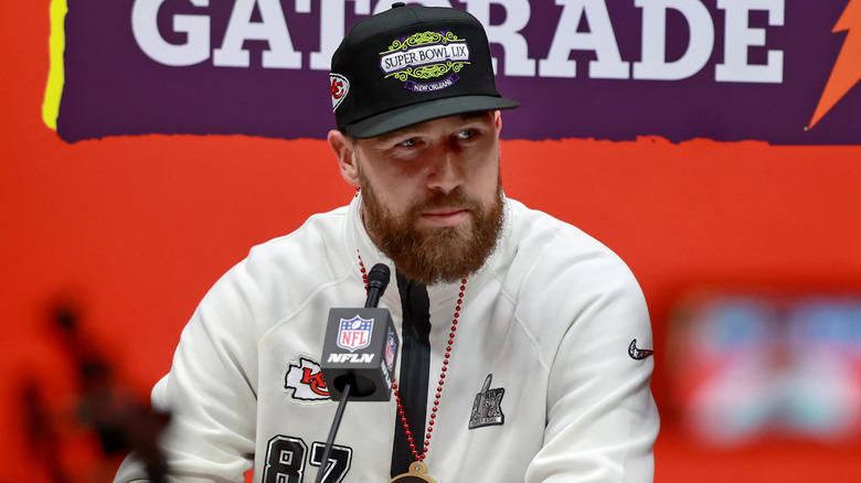 Travis Kelce in a black cap sits at a table with a microphone, cameras surround him.
