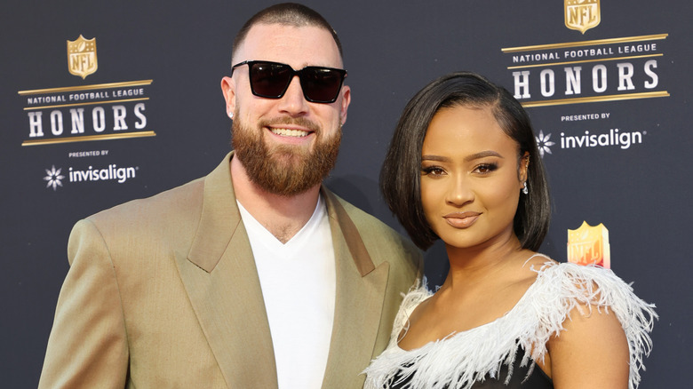 Travis Kelce in a tan suit and Kayla Nicole in a gown with white fringe