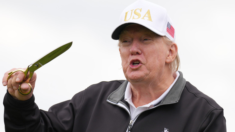 Donald Trump in a white USA hat speaking while holding a giant gold scissors