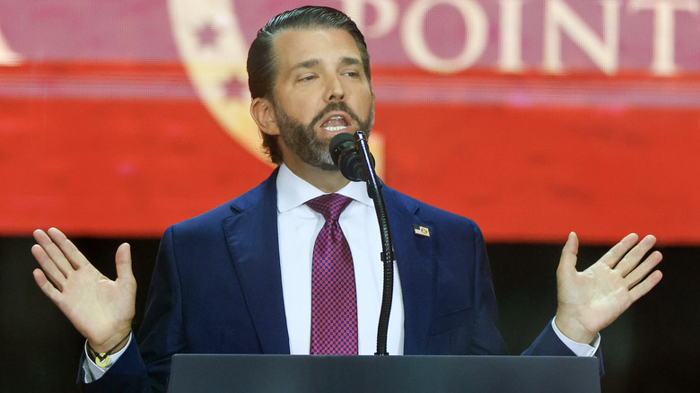 Donald Trump Jr. delivering a speech into mic at podium with hands spread in gesture