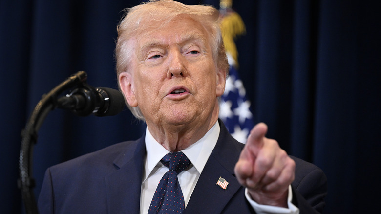 Donald Trump pointing a finger as he speaks in front of a microphone in a black suit with an American flag pin