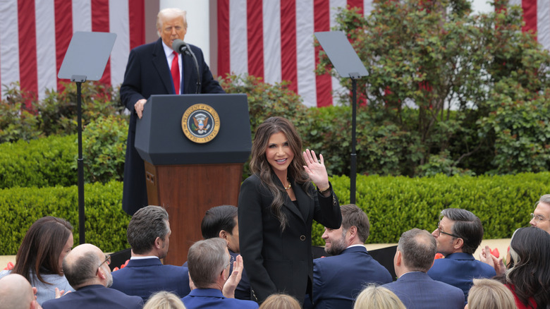 Donald Trump speaking at podium while Kristi Noem waves