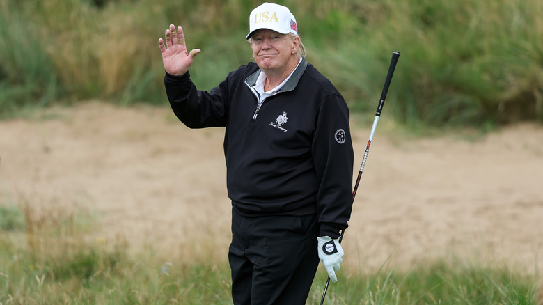 Donald Trump waving to spectators on the golf course wearing a white USA hat