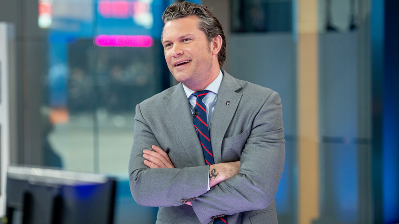 Pete Hegseth with his arms crossed, standing in a studio.