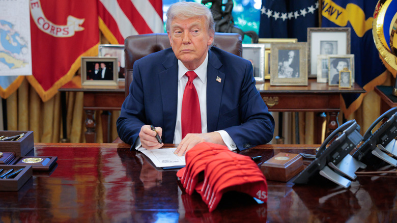 Donald Trump sitting in the Oval Office with a stack of MAGA hats displayed on his desk