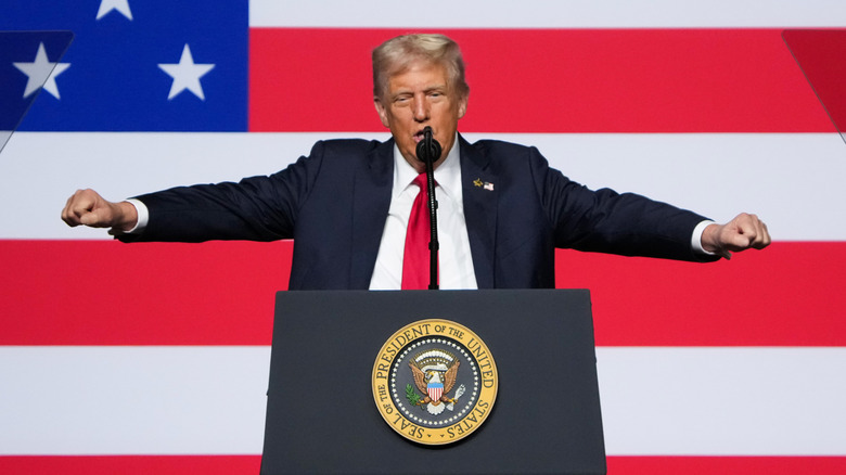 Donald Trump speaking at podium in a suit with his hands raised and an American flag behind him