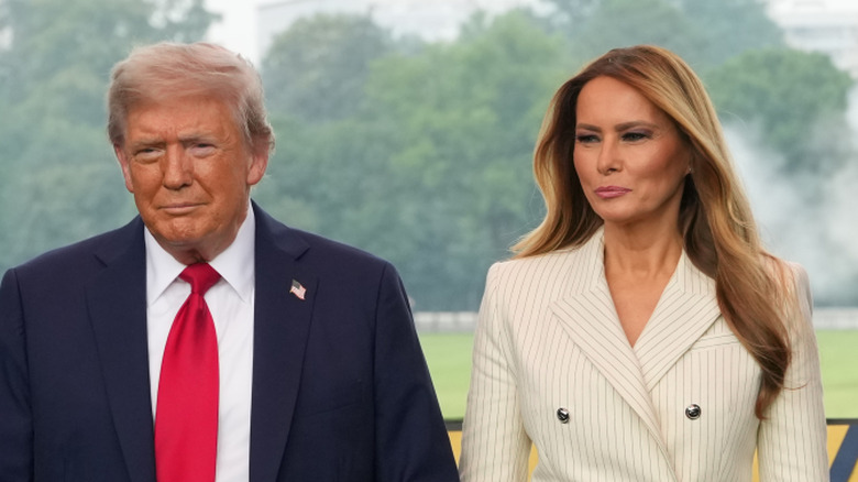 Donald Trump wearing a suit and red tie while holding Melania Trump's hand, who's wearing a white blazer and skirt.