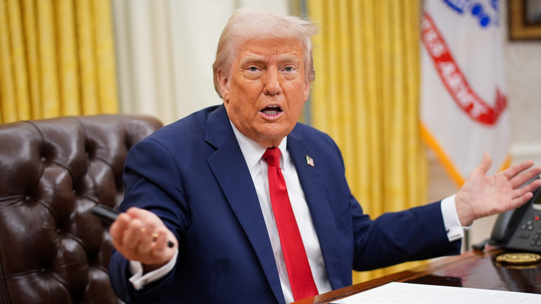 Donald Trump mid-sentence with arms open sitting at his desk in Oval Office
