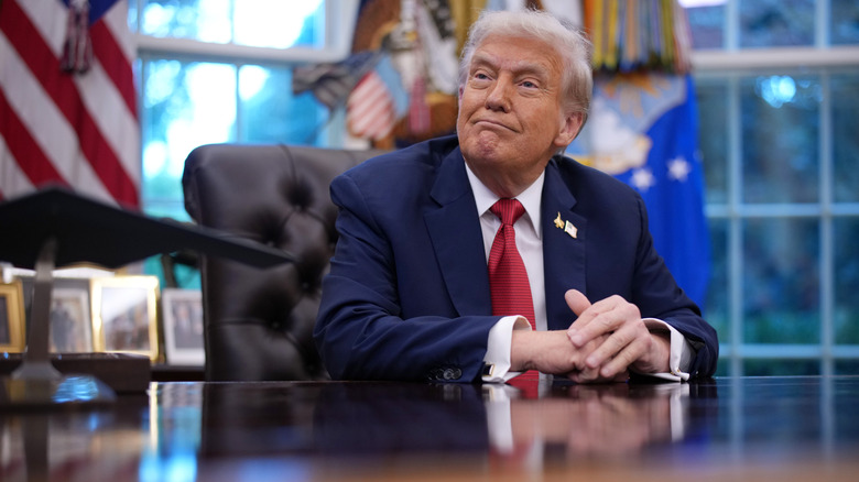 Donald Trump sitting behind the Resolute desk in a navy suit with a red tie