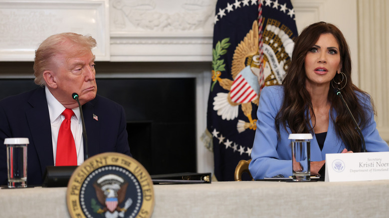 Donald Trump in suit sitting behind table with Kristi Noem to his right in a blue suit.