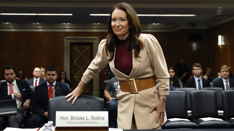 Brooke Rollins looking off as she pulls out a chair to a desk in a dark paneled room