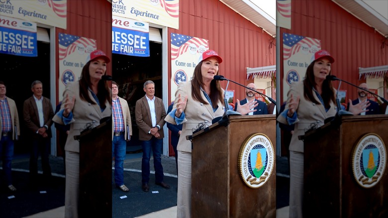 Brooke Rollins in front of a barn, speaking into a microphone with people behind her.