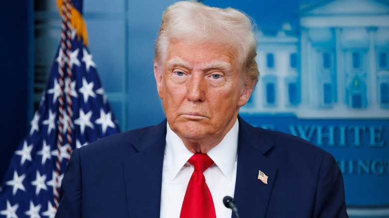 Donald Trump staring off, expressionless, American flag and White House logo in the background