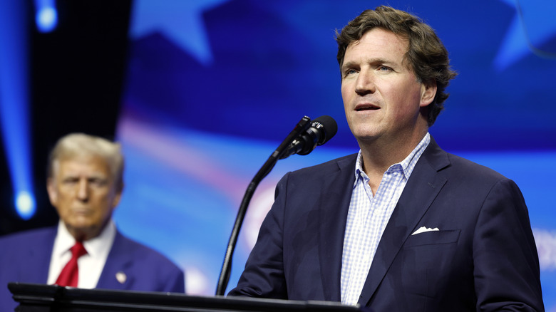 Tucker Carlson addressing a crowd from the podium with Donald Trump out of focus behind him