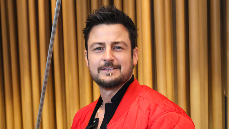 Tyler Hynes wearing a red jacket posing in front of a beige background