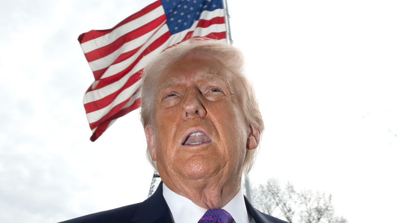 Donald Trump photographed from a low angle with his mouth open and an American flag behind him.