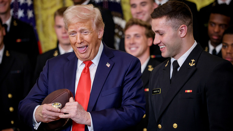 Donald Trump laughs while holding a football and large gold ring as Navy linebacker Colin Ramos smiles beside him.
