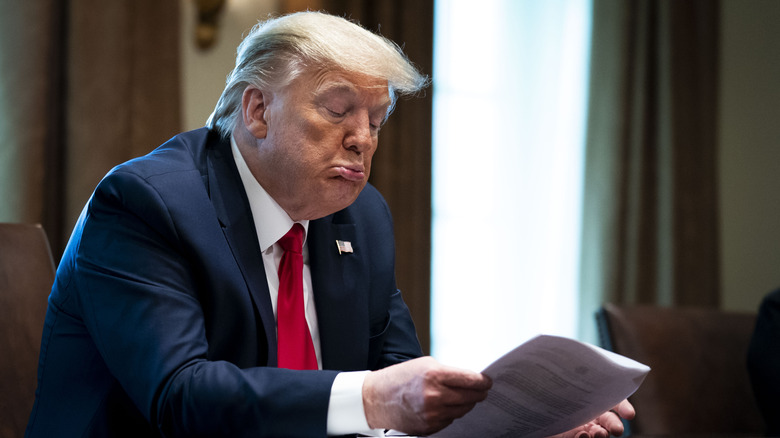 A seated Donald Trump sticks his lips out while reading a document.