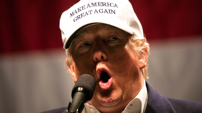 Donald Trump wears a white "Make America Great Again" hat and speaks into a microphone.
