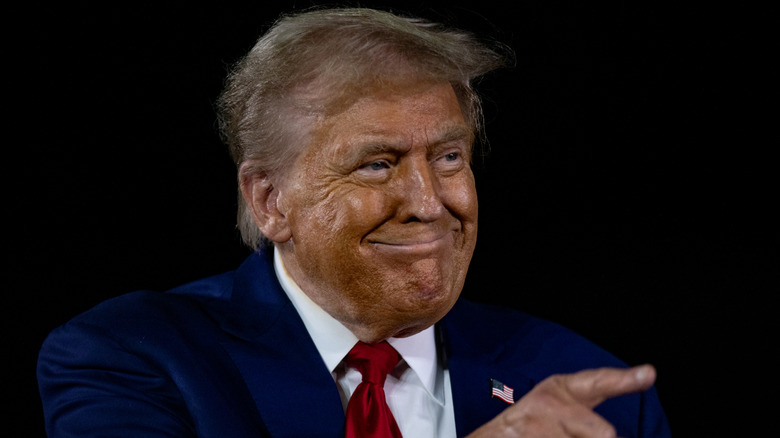 Wearing dark bronzer, Donald Trump smiles with his lips closed and points.