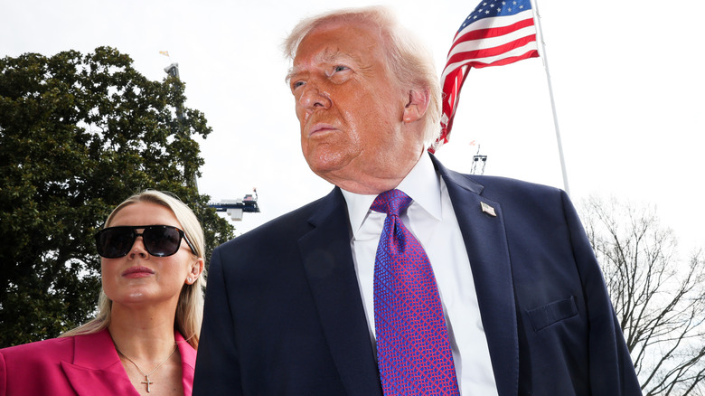 Karoline Leavitt wears a pink blazer and stands behind a sweaty-faced Donald Trump, who wears a purple tie.